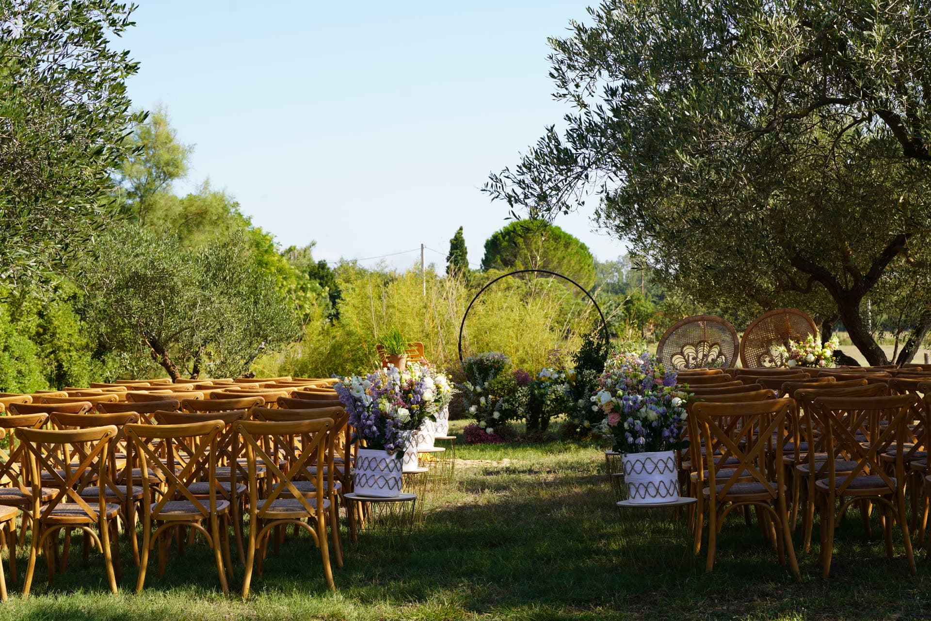 Une cérémonie de mariage dressée sous les oliviers, chaises en bois et compositions florales.