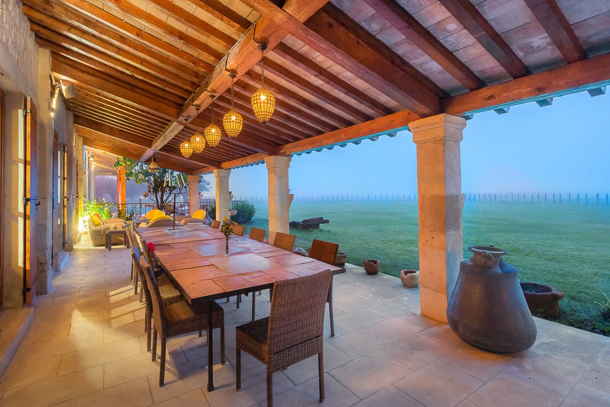 La terrasse couverte dressée pour dîner, charpente en bois et vue sur les prés au crépuscule.